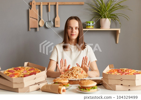 Portrait of serious confident woman with brown hair wearing white casual T-shirt sitting at table in kitchen, keeps diet, showing stop gesture, doesn't eat junk food, prefers healthy eating. 104329499