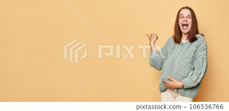 Amazed positive cheerful pregnant woman wearing knitted warm sweater standing isolated over beige background pointing away at copy space for advertisement. 106536766