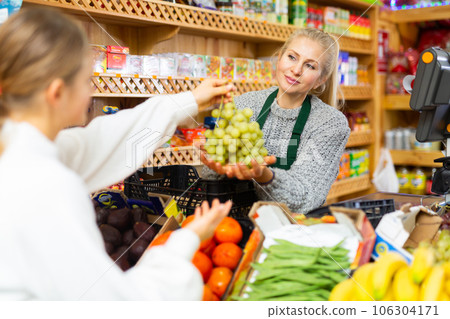 Saleswoman taking grape from female customer for weighing in grocery 106304171