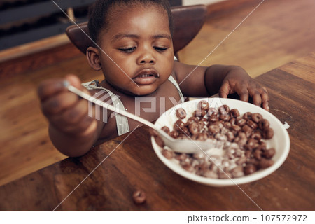 Black child, cereal and eating baby in a home kitchen with food and bowl at breakfast. African girl, nutrition and youth in a house with hungry kid relax with chocolate fiber snack in the morning 107572972