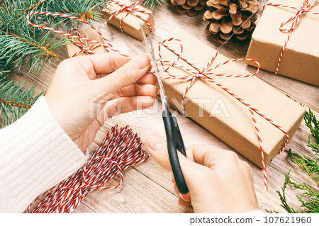 Woman wrapping christmas present , girl prepares xmas gifts with fir tree and pine cone. Hand crafted gift on wooden background with Christmas decor. Top view, copy space. Toned 107621960