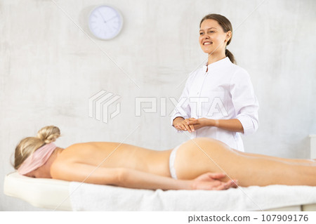 Smiling young female masseur in white coat standing in massage room 107909176