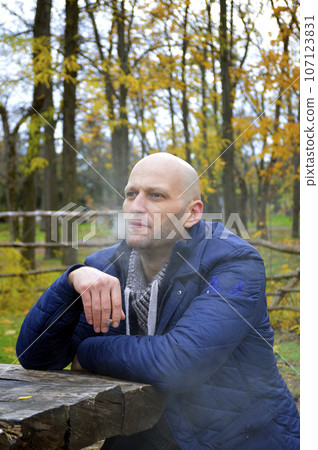 bald young man in a jacket smokes a cigarette on a bench in the autumn park 107123831