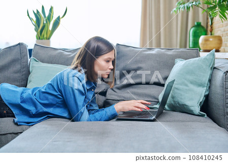 Young woman lying on sofa in living room using laptop 108410245