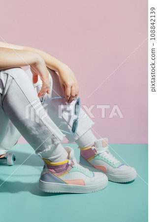 Close up female legs in white jeans and retro style high-top multicolor sport sneakers shoes sitting on the skateboard on turquoise and pink background. Vintage retro fashion style of 80s - 90s vibes. 108842139