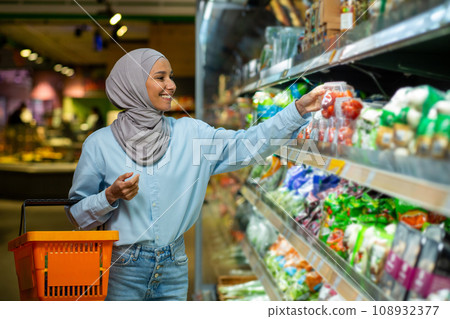 Joyful smiling Arab woman in a hijab chooses products in a supermarket, walks with a basket in her hands along the rows, healthy lifestyle. 108932377