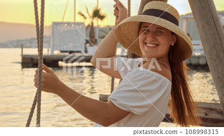 Brunette woman with flowing hair, dressed in a long white gown swaying on a beachside swing at sunset. Tourism, summertime, holiday getaways, and beachfront vacations 108998315