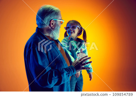 Senior man and little girl, grandfather and granddaughter watching TV over gradient orange background in neon light 109854789