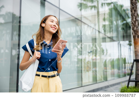 Young Asian woman using smartphone and earbuds to watch social media videos and make virtual calls. This portrait is the epitome of multitasking. 109988787