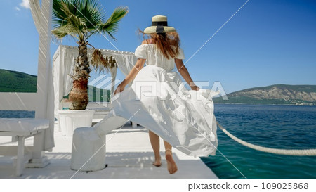 Lady in a flowing light dress and straw hat enjoys a carefree walk on a wooden pier at the beach, evoking the spirit of a summer vacation 109025868