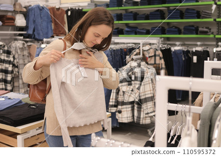 Caucasian woman enjoying shopping in fashionable mall holding stylish attire on hanger looking at model walking in boutique for wardrobe updating 110595372