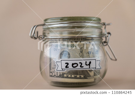 Saving Money In Glass Jar filled with Dollars banknotes. 2024 year transcription in front of jar. Managing personal finances extra income for future insecurity 110864245