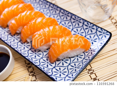 Blue rectangular ceramic plate of delicious salmon nigiri with soy souse on bamboo mat. 110391268