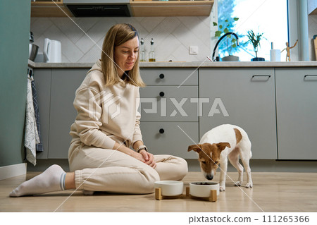 Woman putting food bowl with feed for her dog on the floor in kitchen, Female owner spending time together with pet at home, Animal feeding and pet care 111265366