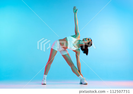 Sportive young woman with slim, fit body in sportswear, doing stretching exercises against blue studio background in neon light 112507731