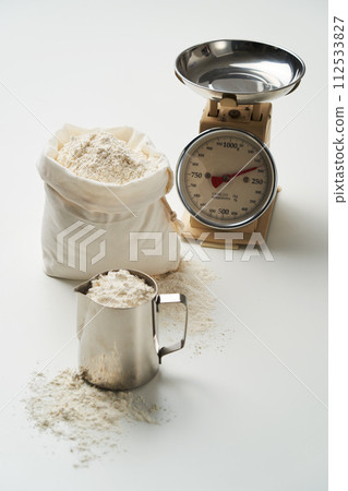 A small bag and measuring cup are filled with flour in front of the scale 112533827