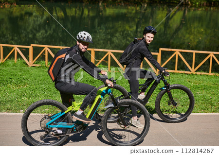 A blissful couple, adorned in professional cycling gear, enjoys a romantic bicycle ride through a park, surrounded by modern natural attractions, radiating love and happiness 112814267