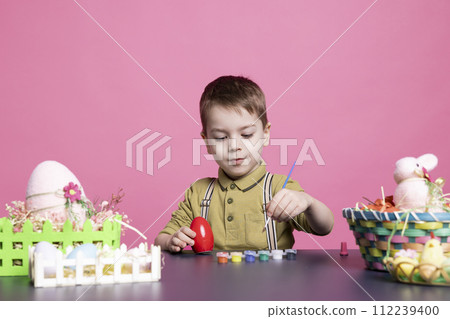 Smiling cute preschooler painting eggs and ornaments for easter holiday preparations, using watercolor and paintbrushes to decorate items. Little happy buy having fun with creativity. 112239400