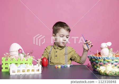 Lovely youngster with a smile crafting eggs and decorations for the Easter festivities, using art supplies and watercolor paint. Little pleased boy feeling excited about painting activity. 112310848