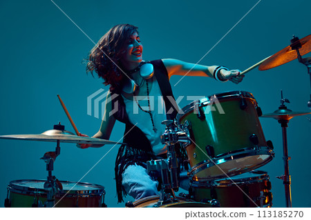 Beautiful, energetic young woman, artistic musician playing drums against cyan background in neon light 113185270