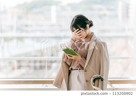 A businesswoman worried while looking at her notebook or schedule book at a station, airport, or public facility (worried, troubled, thinking, busy) 113208902