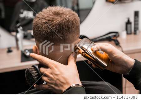 Barber cuts red-haired client occiput with adjustable trimmer in barbershop closeup. Professional master does masculine hairstyle to man in salon 114517393