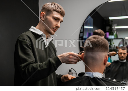 Focused barber cuts client hair on occiput with trimmer in barbershop. Male hairstylist does stylish hairdo to man in beauty salon. Haircut shape 114517457