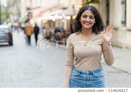 Indian woman smiling friendly at camera, waving hands hello, hi, greeting or goodbye in city street 114626817