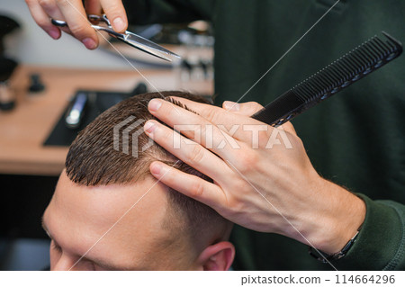 Hairstylist carefully cuts the brunette hair of the client with scissors at the barbershop.  114664296