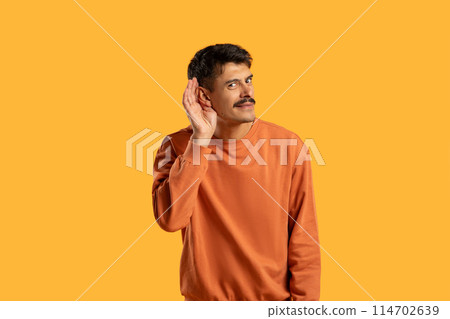 A man wearing an orange shirt is shown in the image, holding his hand up to his ear as if he is listening intently. He appears to be focused on hearing something important. 114702639