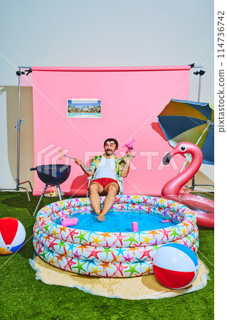 Young man in vibrant outfit with tropical shirt relaxing in inflatable swimming pool and cooking barbeque-sausages. 114736742