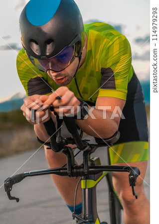 Triathlete riding his bicycle during sunset, preparing for a marathon. The warm colors of the sky provide a beautiful backdrop for his determined and focused effort. 114979298