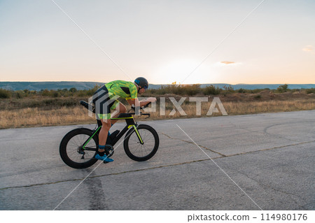 Triathlete riding his bicycle during sunset, preparing for a marathon. The warm colors of the sky provide a beautiful backdrop for his determined and focused effort. 114980176