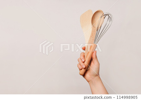 Hand holding a wooden spoon, a wooden spatula, and a whisk against a plain beige background, copy space 114998905