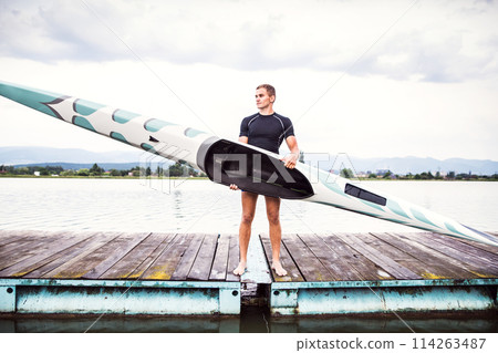 Young canoeist holding canoe and paddle, going into water, wstanding on wooden dock. Concept of canoeing as dynamic and adventurous sport 114263487