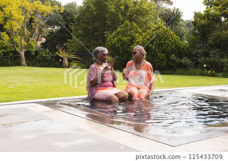 Senior African American woman and senior biracial woman enjoy drinks by the pool 115433709