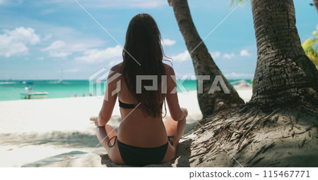 Woman in bikini sitting in lotus pose under palm tree on tropical Boracay island. Long hair brunette enjoy beautiful ocean scene with white sand and turquoise water. Travel, tourism, holiday, relax 115467771