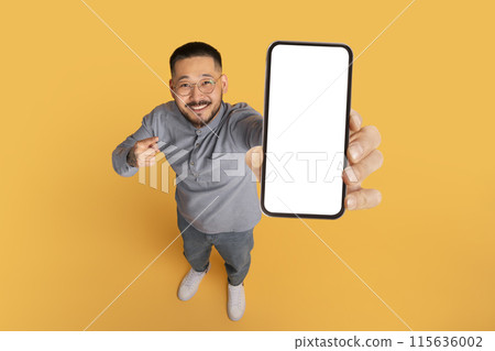 Check This. Smiling Asian Man Pointing At Big Blank Smartphone In His Hand, Happy Millennial Guy Advertising Online Offer Or Mobile App, Standing Over Yellow Studio Background, Collage, Mockup 115636002