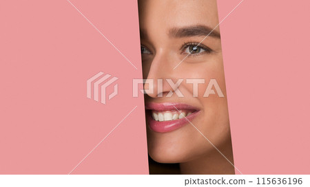 A woman with a bright smile peeks through a pink paper backdrop, showcasing her clear complexion and vibrant expression under studio lighting. 115636196