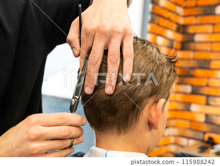 Man barber cutting little boy's hair using comb and scissors. Child getting haircut from adult male, likely barber. Professional hairdresser and cute client at modern barbershop. 115988242