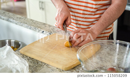 In the contemporary ambiance of a modern kitchen, a young man engages in dinner preparations. His current activity entails meticulously slicing small rainbow potatoes in half on a wooden cutting board 115058209