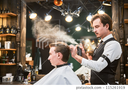 Professional barber using hairspray to finish hairstyle. Stylish hairdresser making final touches. Young customer getting fresh haircut. 115261919