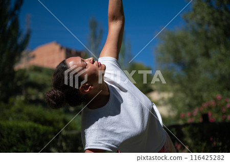 Woman practicing yoga outdoors, stretching arm under blue sky in serene natural setting 116425282