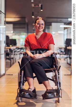 A modern young businesswoman in a wheelchair is surrounded by an inclusive workspace with glass-walled offices, embodying determination and innovation in the business world 116706933