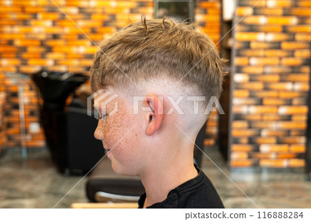 The picture of a small child in a barbershop studio, taken from the side and enjoying the beautiful haircut and the new style, the child has blue eyes and freckles. 116888284