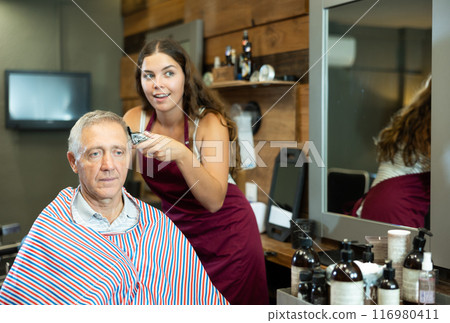 Young female barber making haircut to elderly client in barbershop 116980411