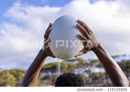 African American young male athlete, on field outdoors, hands holding rugby ball high against a sky 116135888