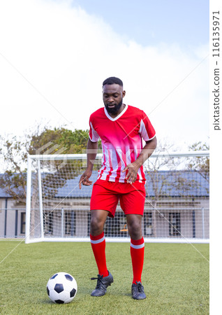 An African American young male athlete wearing red and white soccer uniform dribbling ball on field 116135971