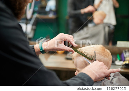 Man barber cutting little boy's hair using comb and scissors. Child getting haircut from adult male, likely barber. Professional hairdresser and cute client at modern barbershop. 116277925