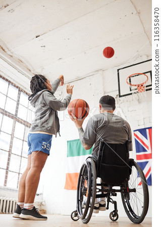 Vertical back view of sportsman using wheelchair in basketball practice with female coach throwing ball in hoop 116358470